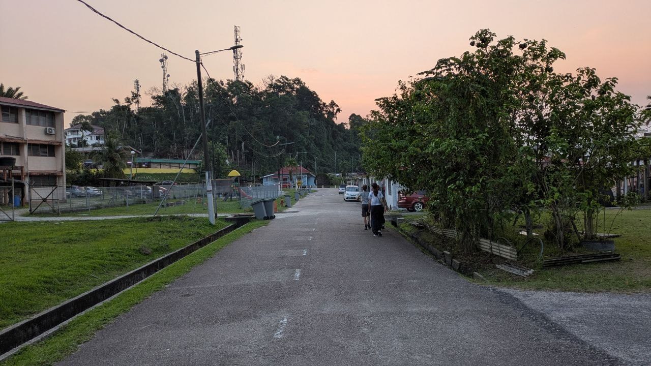A surfaced residential street at dusk, a fenced playground on the left, terraced houses on the right, two people walking along the road.