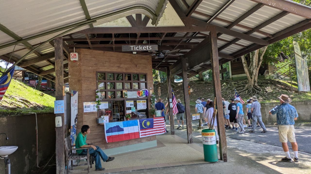 An open wooden hut under a corrugated roof with a Tickets sign above the counter window and a lit OPEN sign, a Malaysian flag in front; on the right a group of visitors walking up the path to the entrance.
