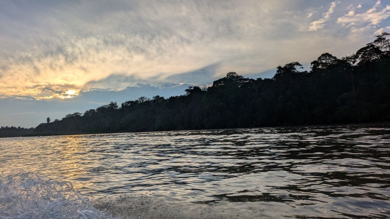 A wide view across the Kinabatangan into the low sun behind a bank of cloud; the dark treeline stands as a silhouette on the right, the boat's spray bottom left.