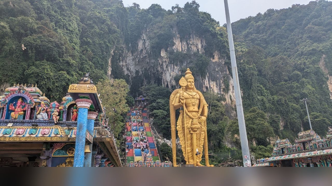The gold Murugan statue in front of the overgrown limestone wall of Batu Caves, the colourful gopuram of a temple on the left, the rainbow staircase in the middle.
