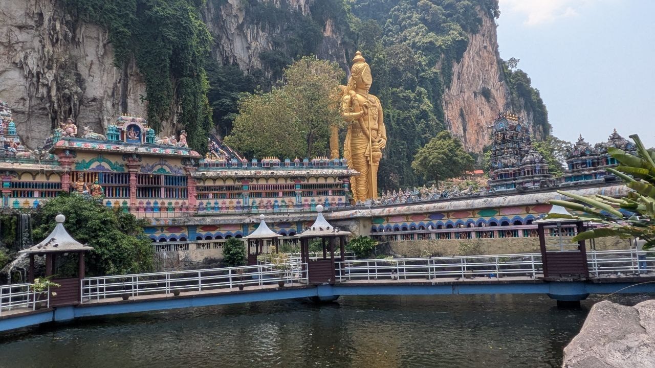 The same statue seen from the pond side, with a long brightly painted row of temple frontages and a blue footbridge over green water in front of it.