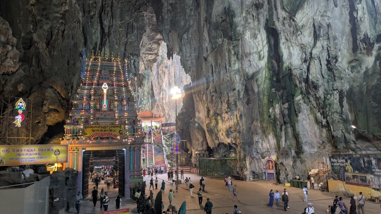 Inside the temple cave at Batu Caves: a colourful temple tower on the left, a high rock face on the right, a shaft of light falling through an opening in the ceiling between them.