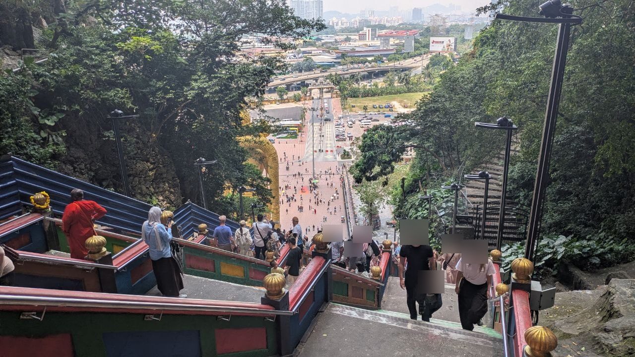 View from the topmost step out over the rainbow-coloured railings, down onto the forecourt, with car parks, a motorway ramp and the high-rises of Kuala Lumpur in the haze behind.