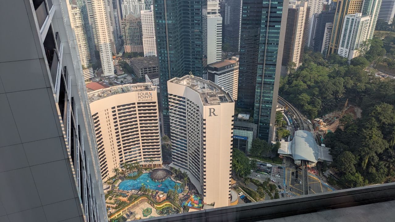 A steep view down onto two hotel towers with a turquoise pool landscape, on the right a station with a curved roof and an elevated track on pillars.
