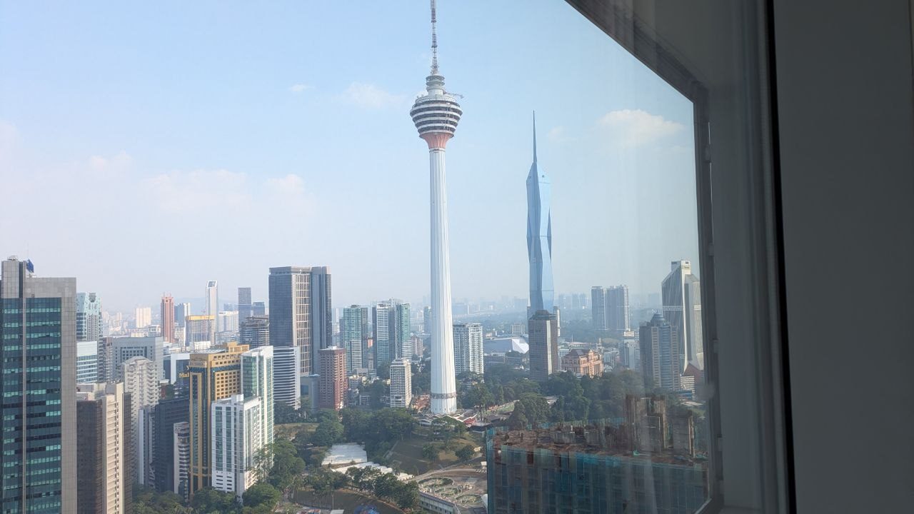 View through a window across the rooftops of Kuala Lumpur: Menara Kuala Lumpur in the centre, the slender Merdeka 118 to its right, densely packed high-rises in front.