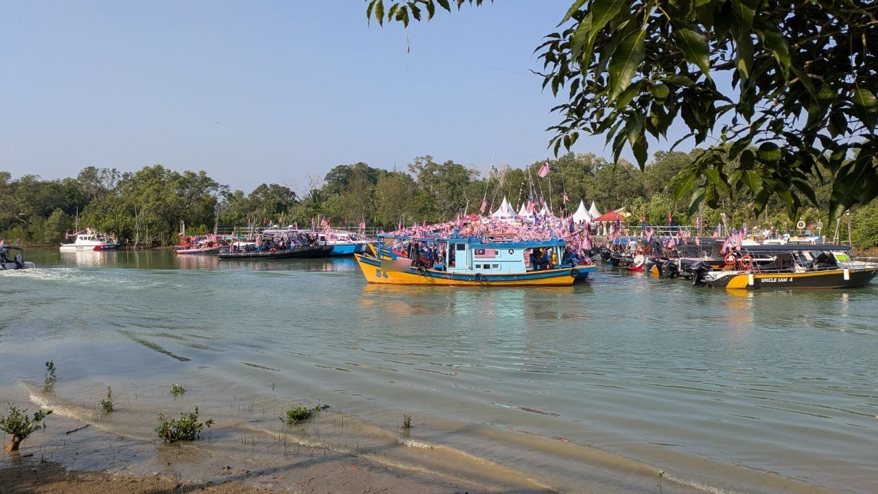 A river harbour with mangroves along the bank: a blue and yellow fishing boat with a Malaysian flag on the wheelhouse, behind it rows of further boats covered in flags.