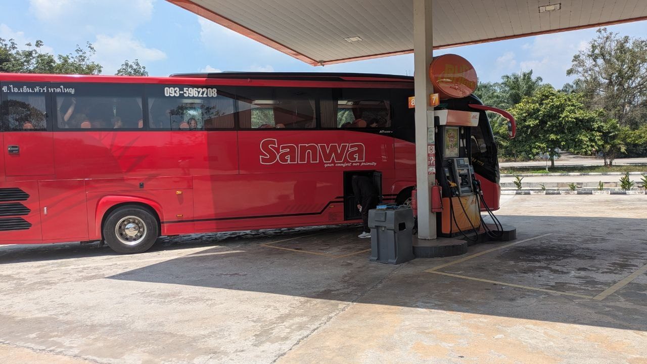 A red coach lettered "Sanwa" refuelling at a petrol station, above it in Thai script the name of an operator from Hat Yai; the luggage hold stands open.