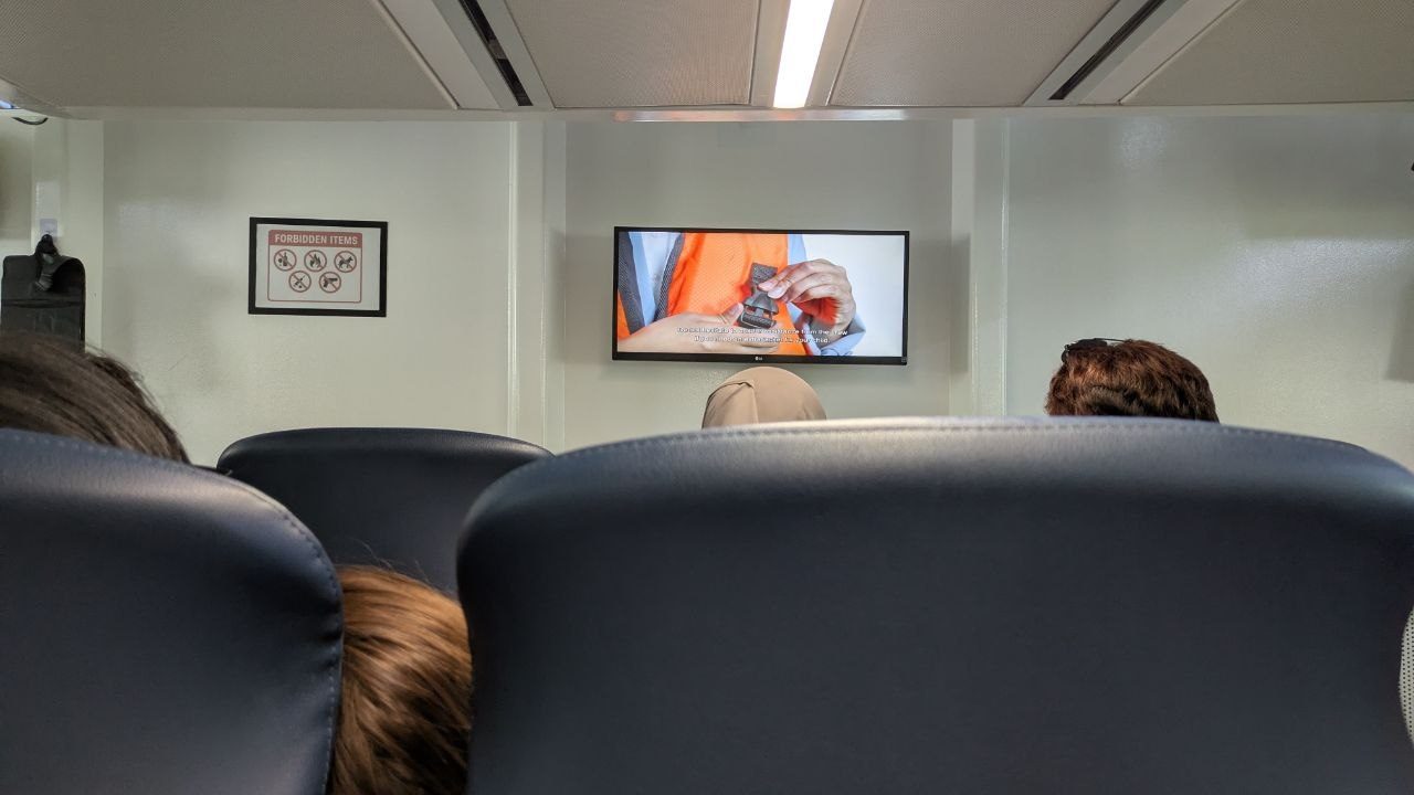View forward inside the ferry cabin: airline-style rows of seats, a screen showing the safety video, next to it a sign listing forbidden items.