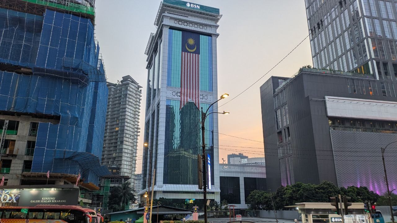 A white bank tower with an outsized Malaysian flag hanging down its glass facade; building sites with blue netting around it and an evening sky.