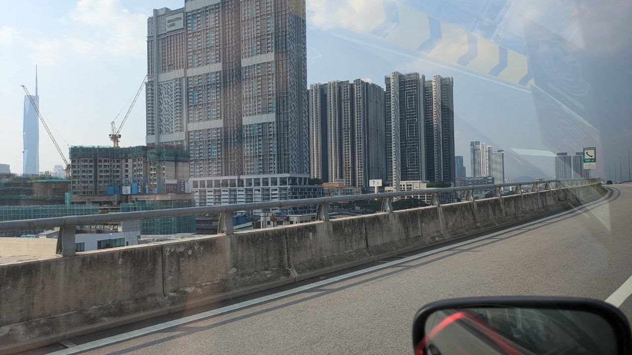 View from a moving car onto a concrete elevated road; behind it on the left residential high-rises and building shells with cranes, far back in the haze the slim Merdeka 118 tower.