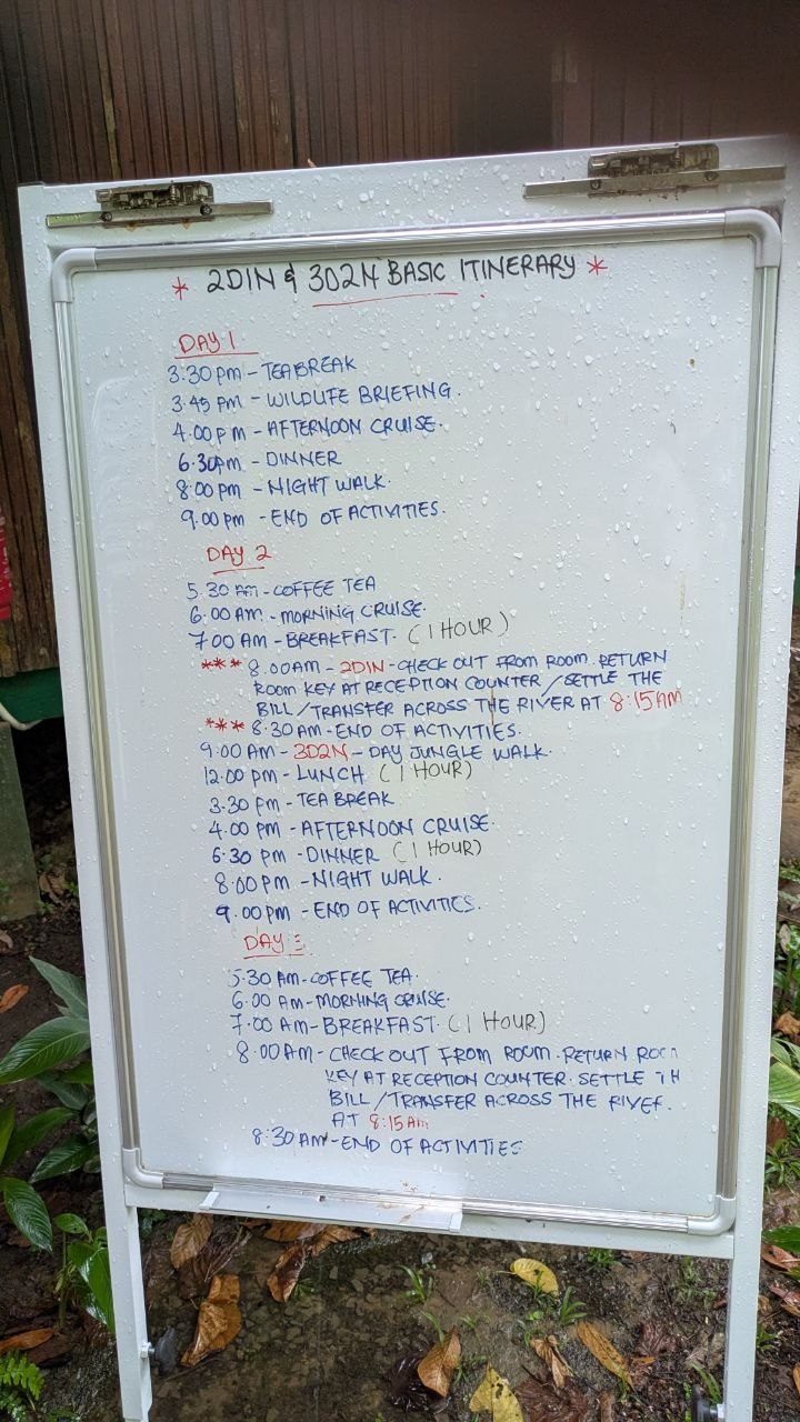 A white outdoor whiteboard under a canopy, handwritten in red and blue with the heading 2D1N and 3D2N Basic Itinerary and the schedules for days 1 to 3; raindrops stand on the board, wet leaf litter at the bottom of the picture.