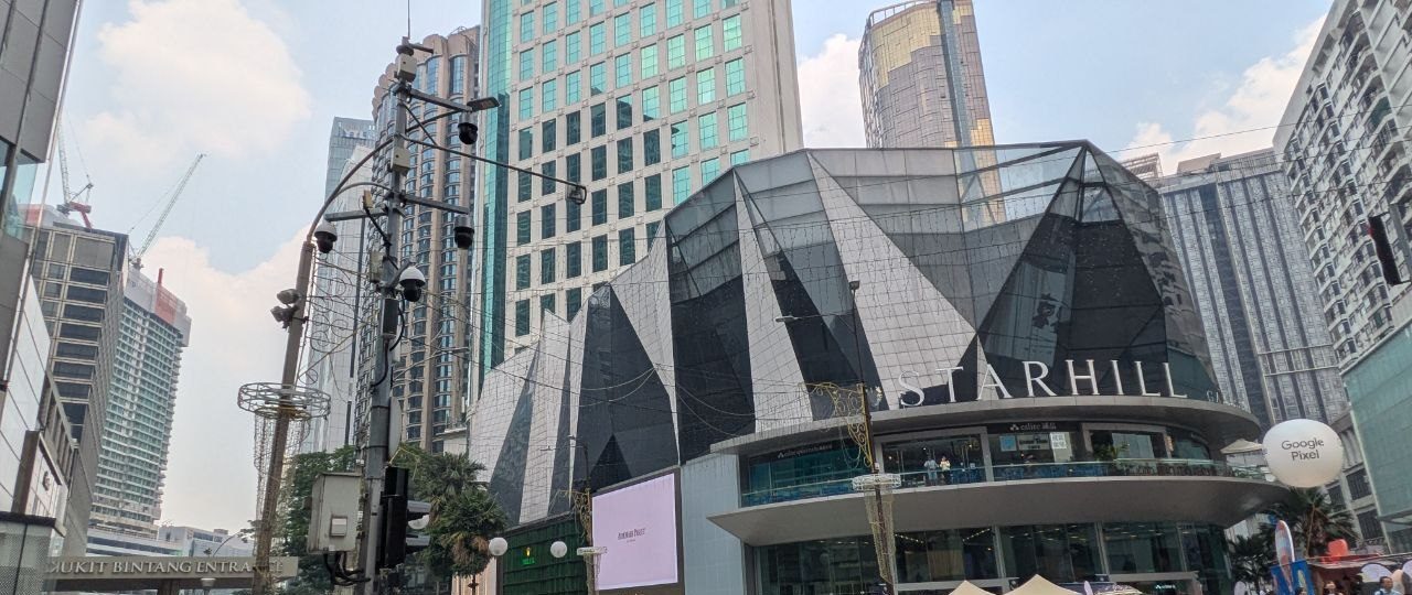 The faceted facade of Starhill Gallery above the junction, on the left the gate reading Bukit Bintang Entrance, on the right the curve of the mall with stalls in front of it.