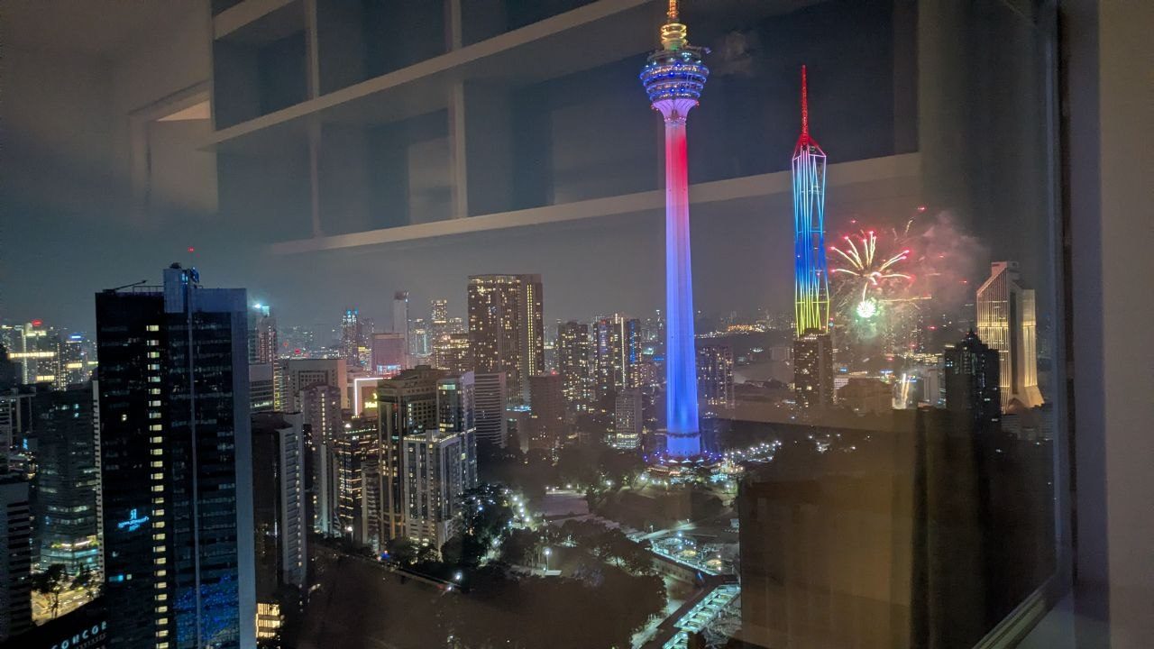 Night view over Kuala Lumpur: the telecoms tower lit in red, white and blue, Merdeka 118 to its right in blue, yellow and red, fireworks above.