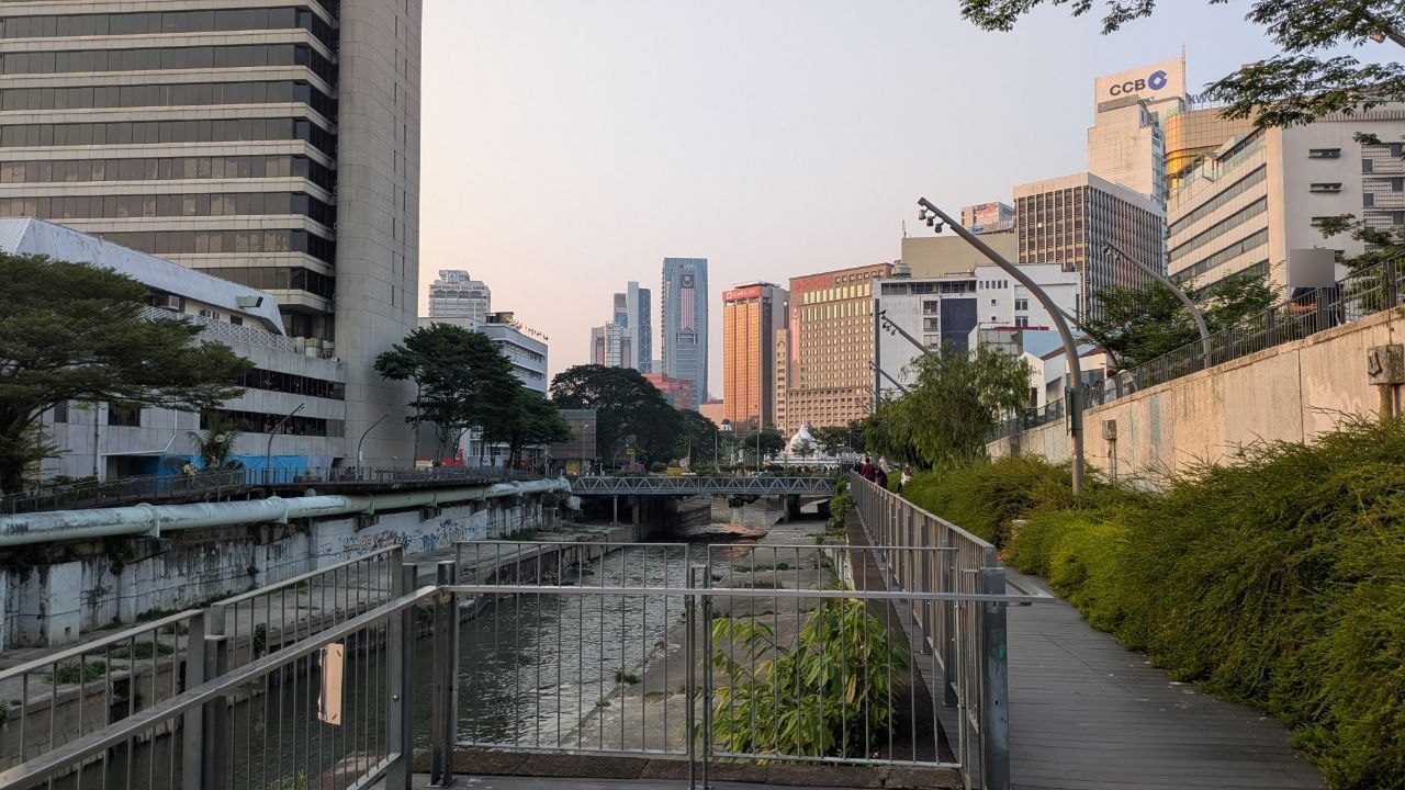 A narrow wooden walkway with a metal railing along the bank of a concrete river bed, densely planted; ahead a blue steel bridge, behind it high-rises and a white dome.