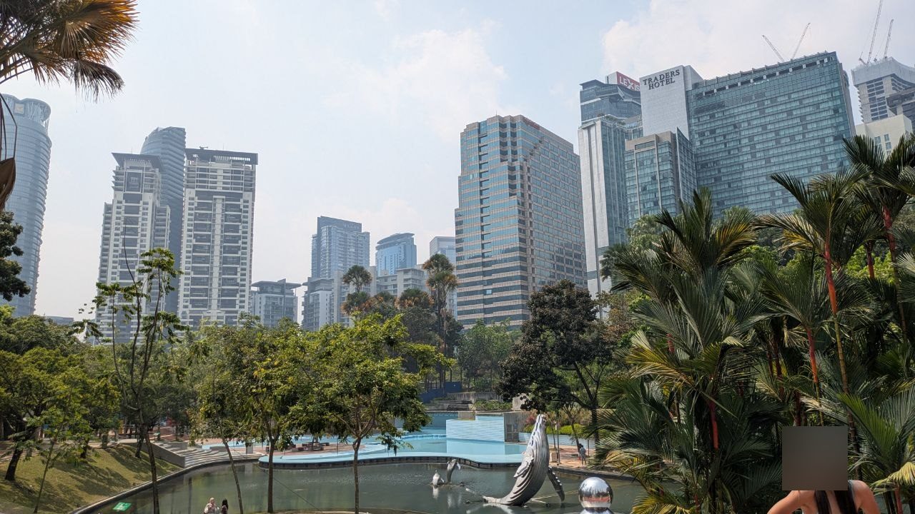 The pond in the park behind Suria KLCC: a humpback whale sculpture standing upright in the water, dolphins and a mirrored sphere beside it; behind them a shallow pale-blue basin, palms and the high-rises of the district.