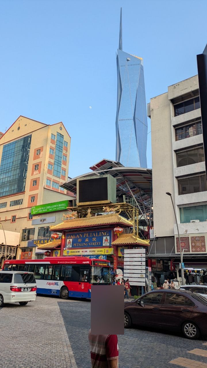 The Chinese gate to Petaling Street with red lettering and lanterns, the market hall behind it and the pointed glass tower of Merdeka 118 above.