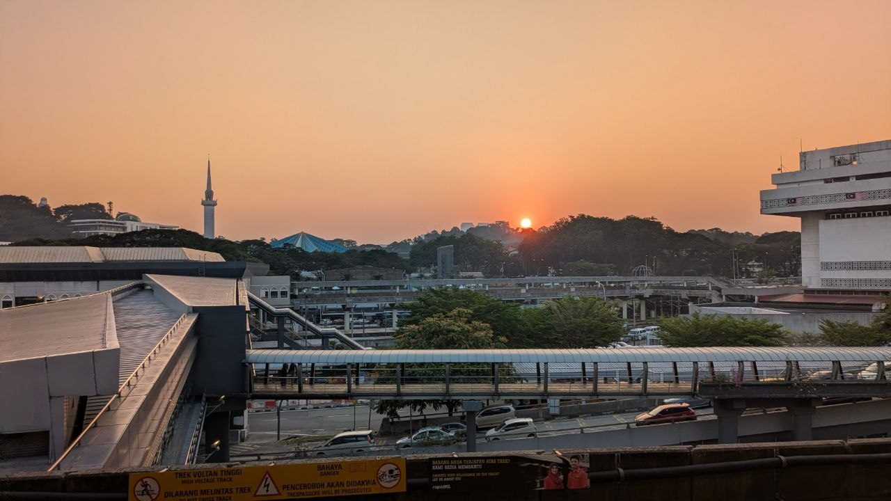 Sunset over Kuala Lumpur from the platform of an elevated station: the sun just above wooded hills, a minaret on the left, road and tracks below.