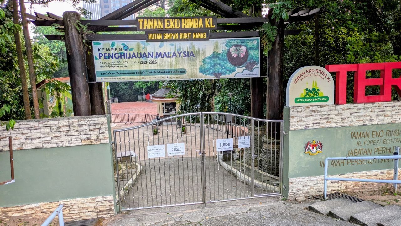 The locked steel gate under the wooden arch reading Taman Eko Rimba KL, Hutan Simpan Bukit Nanas; two signs reading Closed on the leaves, the forest path behind.