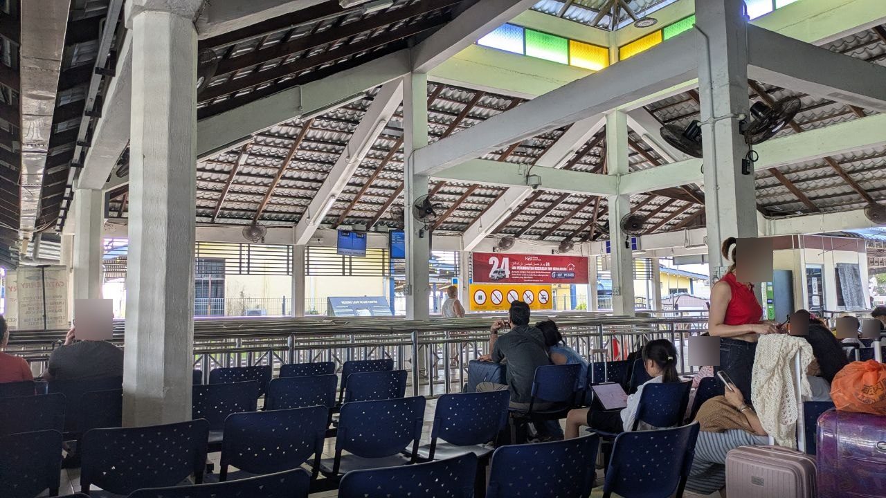 The open waiting hall of the Mersing ferry terminal: tiled roof on posts, ceiling fans, rows of blue seats, waiting passengers.