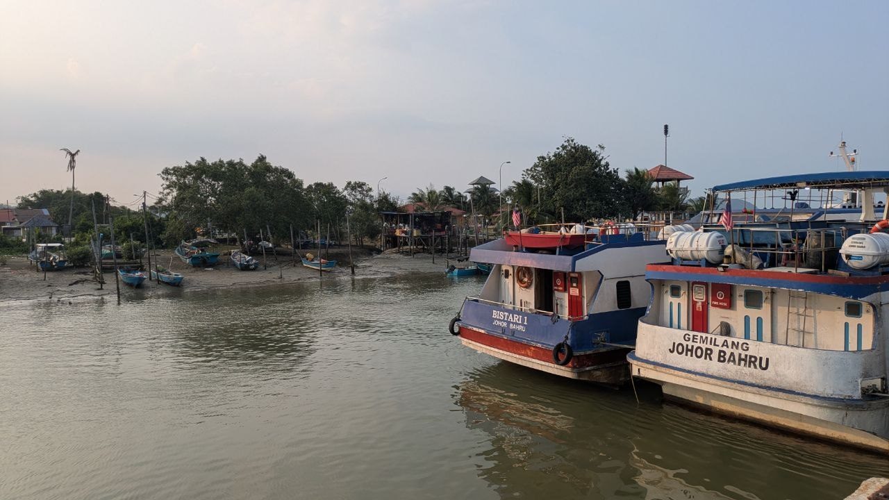 Mersing harbour at low water: small boats lying in the mud along the bank, two larger boats still afloat at the pontoon.