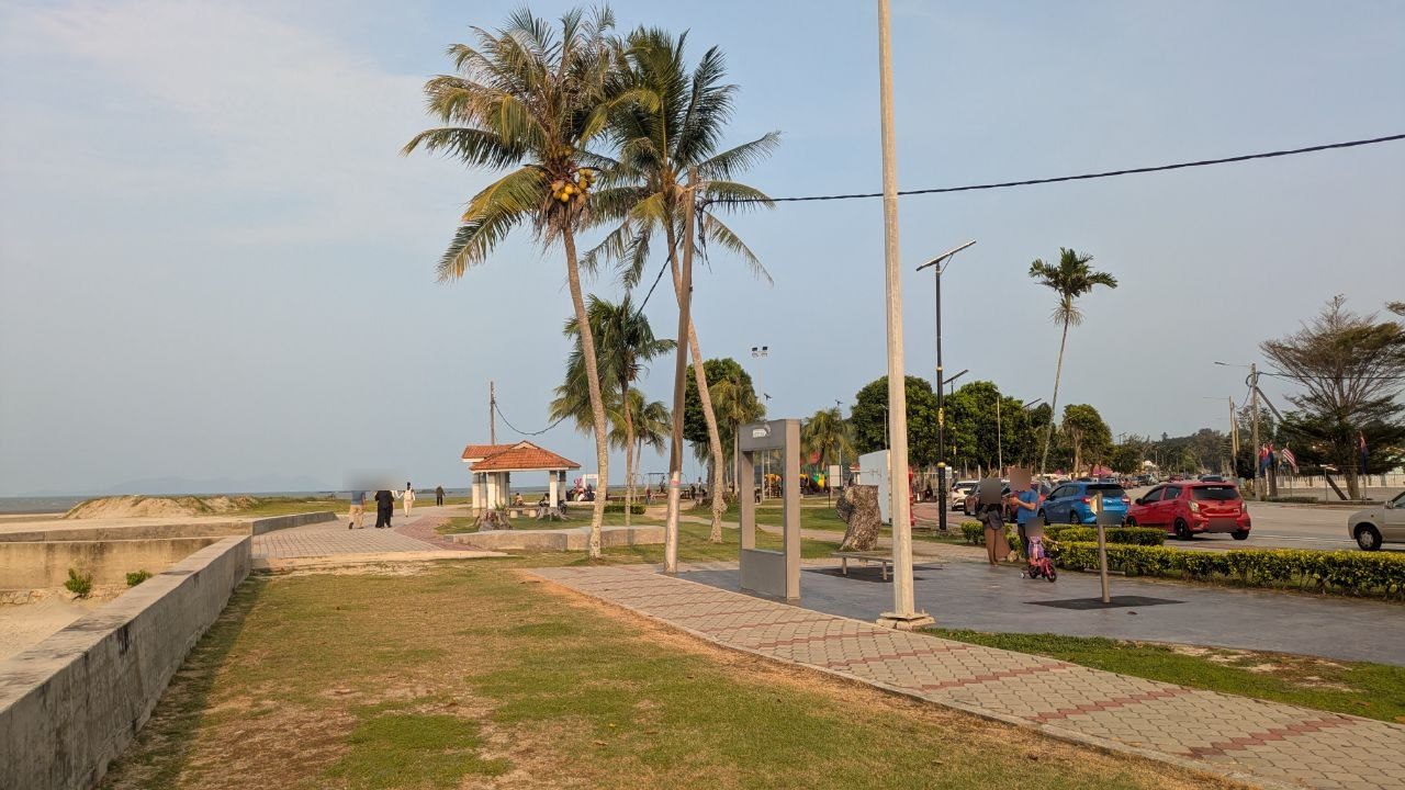The seafront promenade in Mersing in the late afternoon: coconut palms, a paved path, pavilions and the road behind.