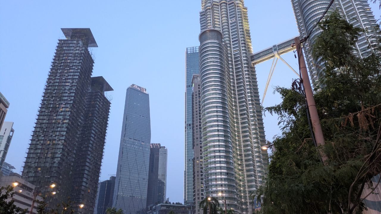The Petronas Towers seen from below at dusk with the skybridge between them; a power pylon and tree branches in the foreground.