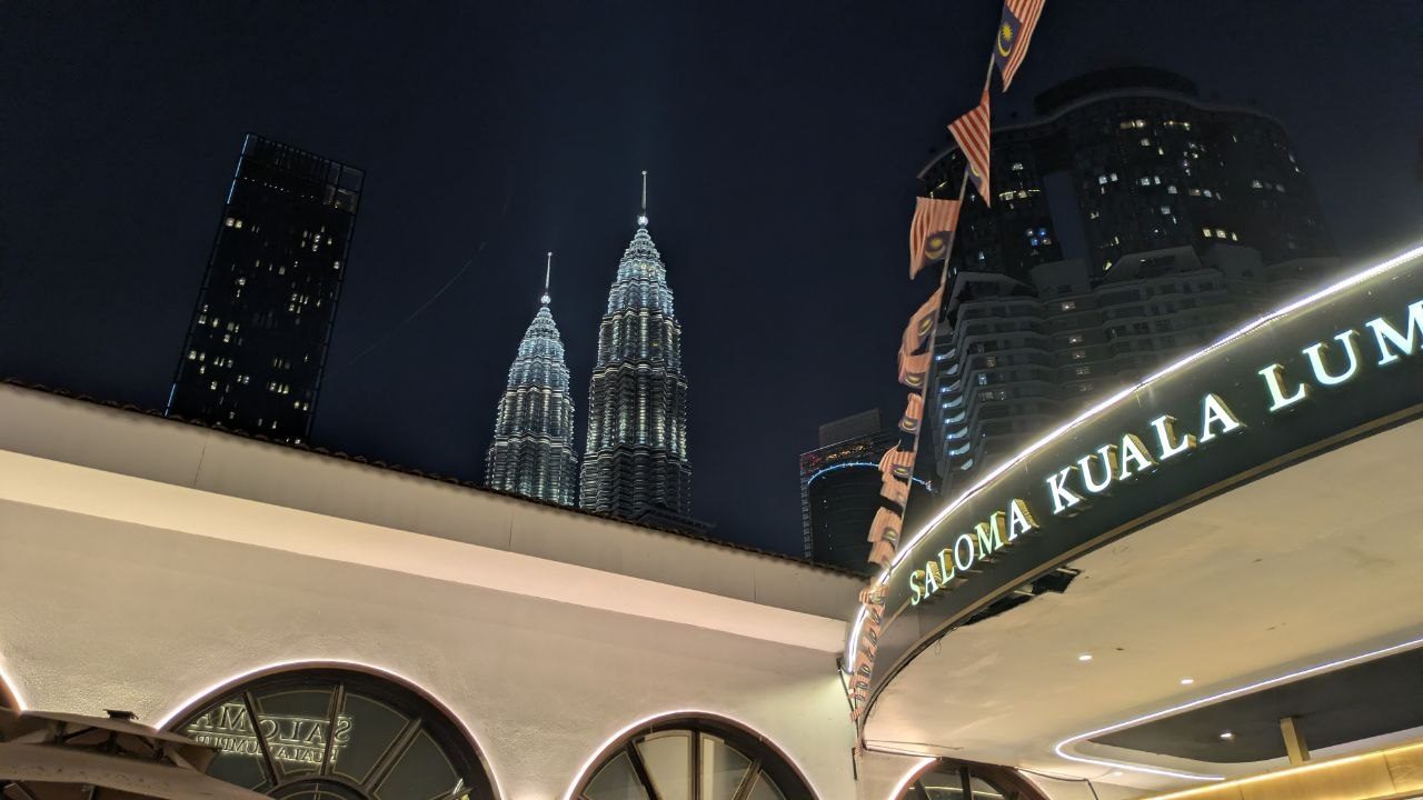 Night shot: an illuminated "Saloma Kuala Lumpur" sign on a curved canopy, Malaysian flags on angled poles in front, the floodlit Petronas Towers behind.