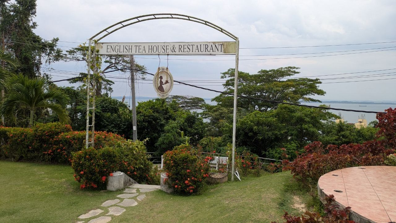 A wrought-iron arch with the lettering English Tea House & Restaurant and an oval sign showing the silhouette of a waitress; behind it lawn, shrubs with red flowers and Sandakan Bay.