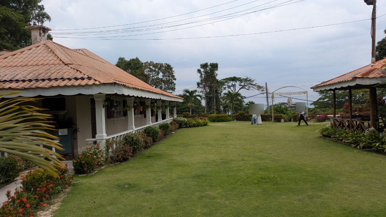 The white-painted main house of the English Tea House with its long veranda under a tiled roof, a wide lawn in front, an open gazebo to the right, the arch and the sea in the background.
