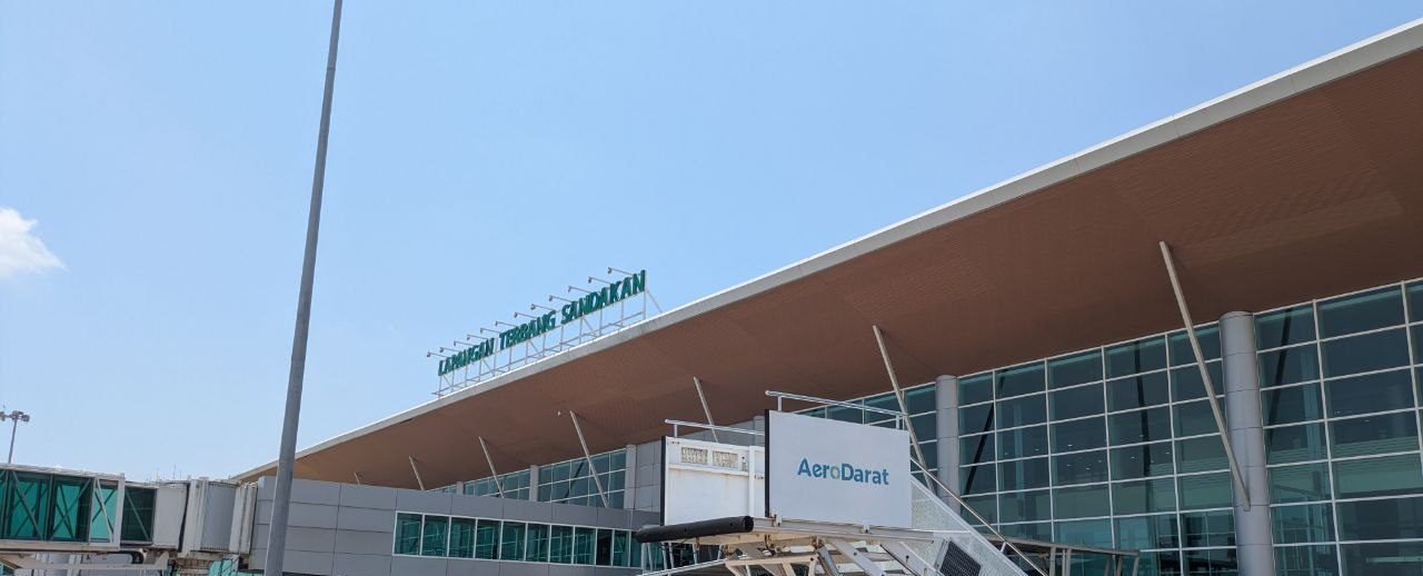 The terminal building at Sandakan airport seen from the apron: a wide brown canopy above a glass front, the name Lapangan Terbang Sandakan in green letters along the roof edge, a sign reading AeroDarat in front of it.