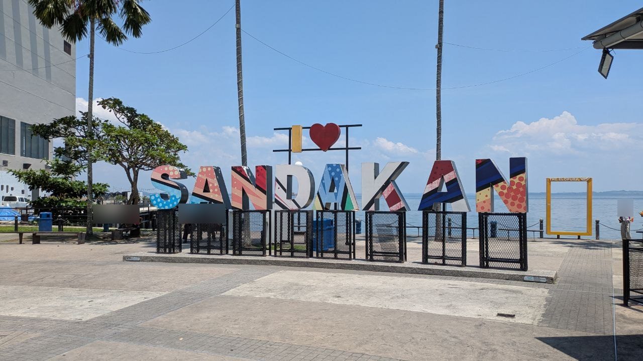 The colourful SANDAKAN lettering with a red heart above it on the harbour promenade, a row of black steel gates in front, the bay behind; to the right a yellow picture frame reading The Elopura.