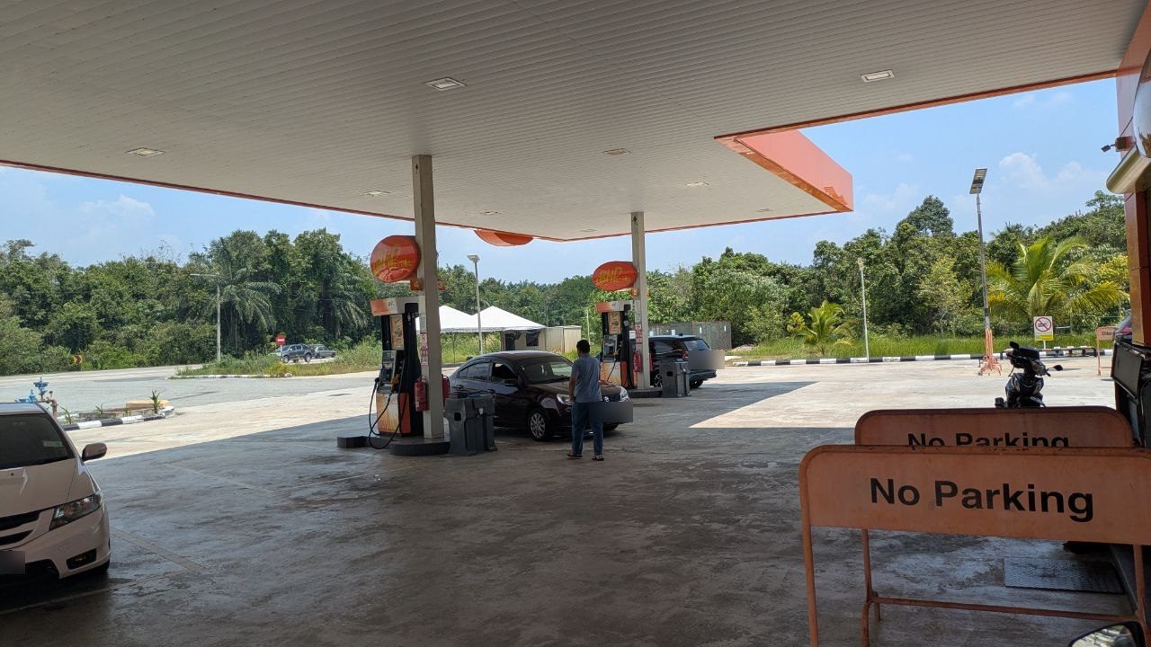 The forecourt of a petrol station under a white canopy, forest and oil palms behind it, on the right an orange barrier reading "No Parking".