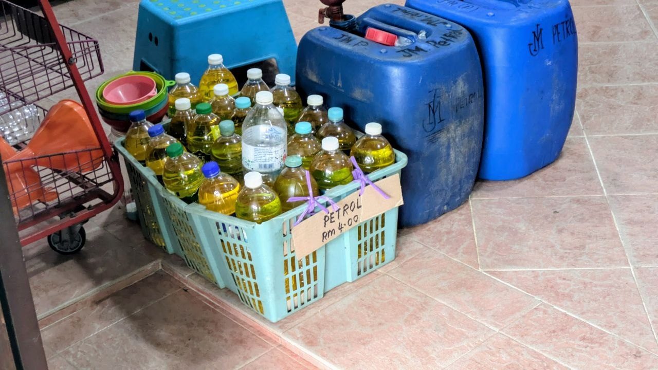 A turquoise plastic crate on a tiled floor, filled with used plastic bottles of yellowish petrol; in front a hand-lettered cardboard sign reading "PETROL RM 4.00", behind it three blue jerrycans stencilled "JM PETROL".