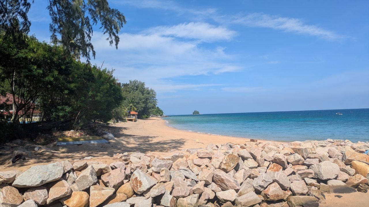 View across a bank of pale boulders onto the empty sandy beach of the bay, trees and the red roofs of the accommodation on the left, turquoise water on the right and a small wooded rock island far out.