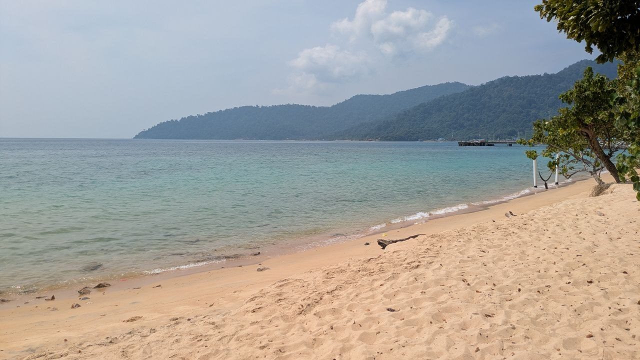 A bright sandy beach with calm turquoise water, forested hills behind it, and a long jetty at the end of the bay.