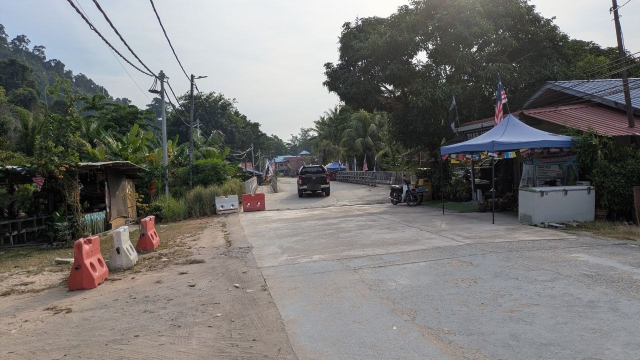 Looking down the village road towards the bridge with its white railing, a blue gazebo with a Malaysian flag on the right, shops and banana plants on the left.