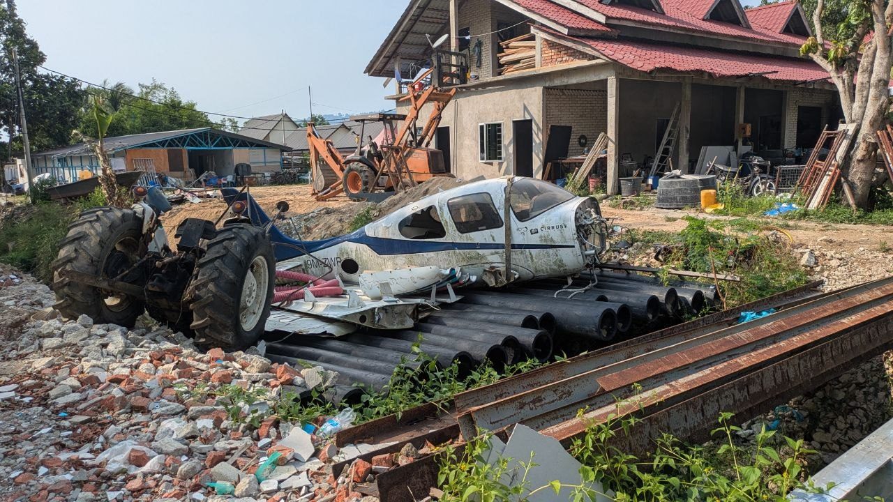 A dismantled white and blue light aircraft marked Cirrus resting on a stack of black plastic pipes and steel beams, a backhoe loader beside it and a red-tiled shell of a house behind.