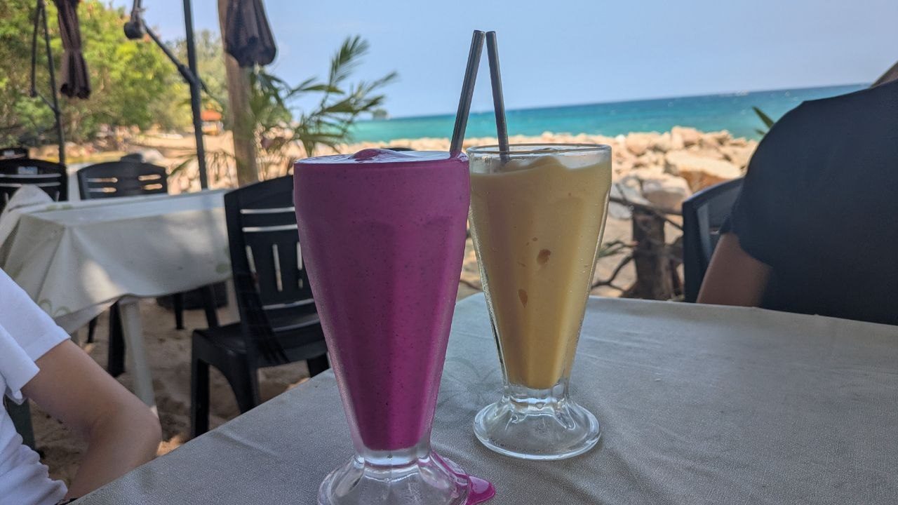 Two tall glasses of fruit drinks on a table by the beach, magenta dragon fruit on the left, pale yellow on the right; behind them boulders, the turquoise water of the bay and palm trees.