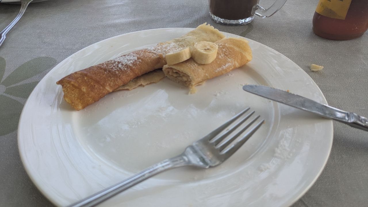 A rolled pancake with icing sugar and three slices of banana on a white plate, knife and fork beside it, behind it a glass mug of coffee and a bottle of syrup.
