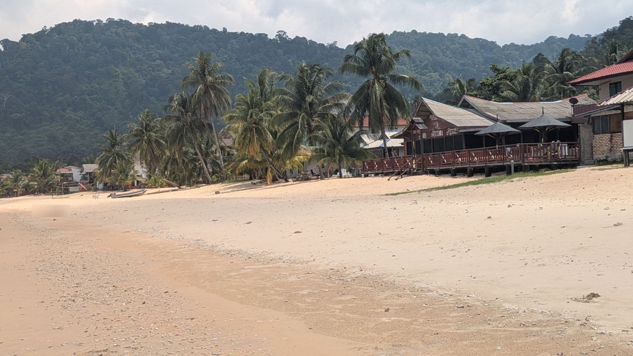 The line of palms on Juara beach, a long restaurant with a wooden terrace and parasols behind them, boats lying in the sand.
