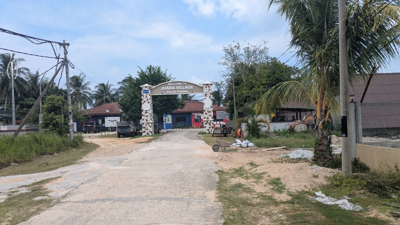 A masonry arch reading "Welcome to Juara Village, Pulau Tioman" over a concrete road, blue and red painted houses with red tiled roofs behind it.