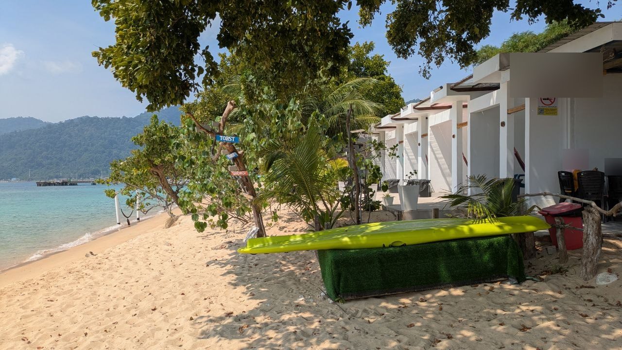 A row of white beach cabins under palms and trees, a kayak on the sand in front of them, the shallow turquoise water of the bay on the left.
