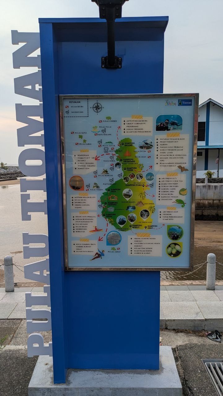 Blue information board on the waterfront path reading "Pulau Tioman"; inside it a drawn island map with villages, dive sites and activity lists.