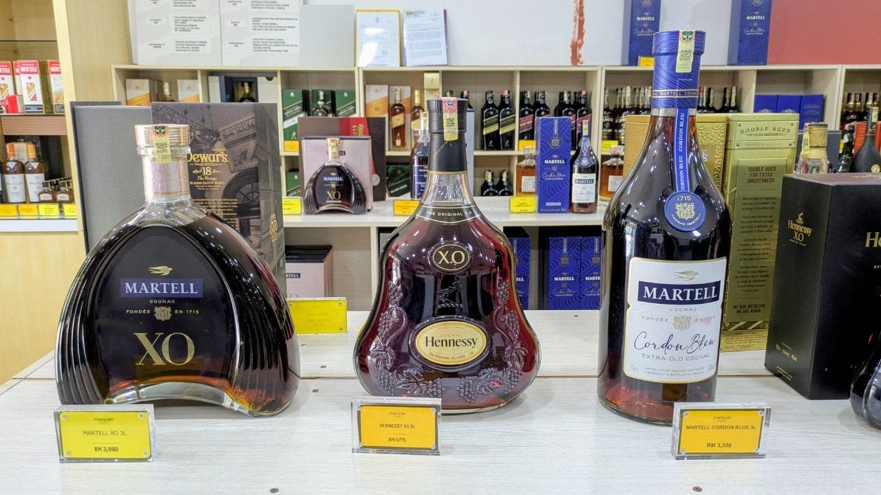 Three three-litre cognac bottles on a shop counter — Martell XO, Hennessy XO and Martell Cordon Bleu — with yellow price tags in front and shelves full of spirits behind.